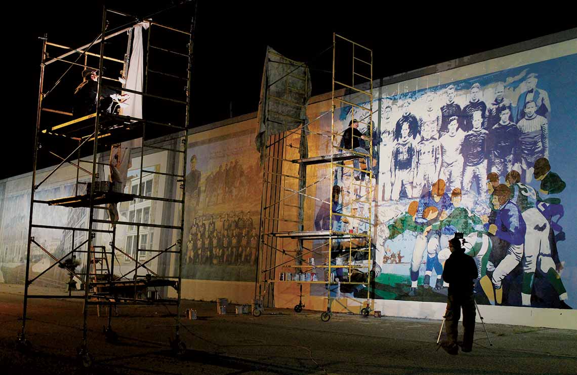 Artists working on a mural at night using scaffolding and large studio lights.